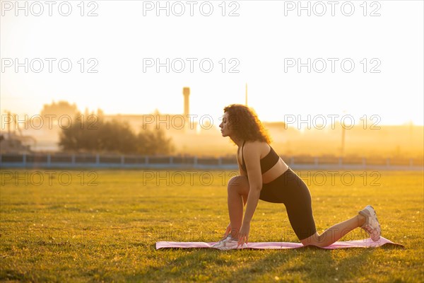 Young curly athletic girl in sportswear doing stretching exercise outdoors on the grass during sunset