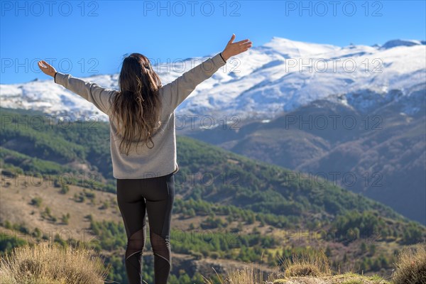 A woman stands with arms raised facing a mountain landscape with a snowy peak under a clear sky
