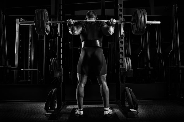 Professional athlete is standing with a barbell on his shoulders and is about to sit down with her. View from the back