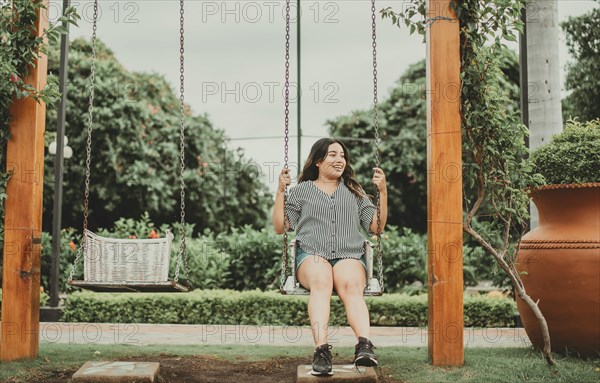 Happy young woman sitting on a swing in a beautiful garden. Portrait of smiling girl sitting on a swing in a garden