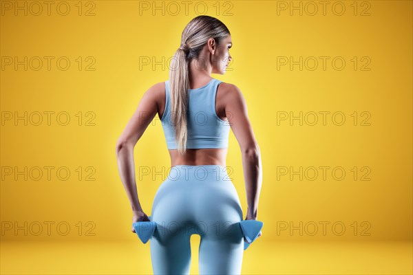 Slender athletic girl in a blue tights posing with dumbbells. Back view.