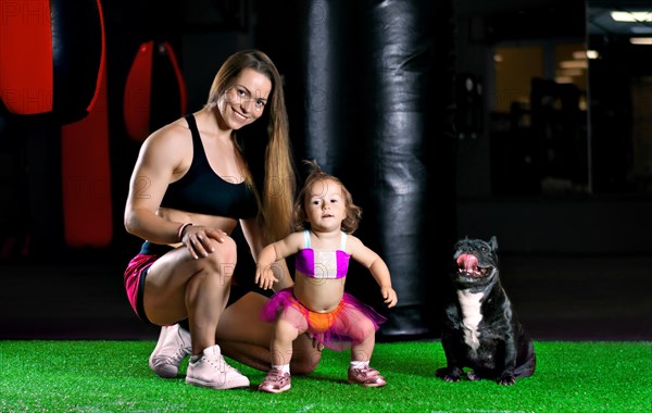 Charming sports mom trains in the gym with her little daughter and a French bulldog.