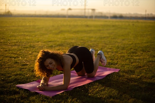 Young curly athletic girl in sportswear performs an exercise on a yoga mat outdoors on the grass during sunset