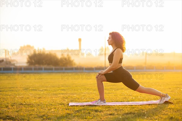 Young curly athletic girl in sportswear doing lunges exercise outdoors on the grass during sunset