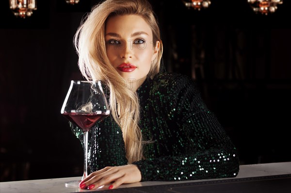 Seductive blonde in a green evening dress posing with a glass of red wine.