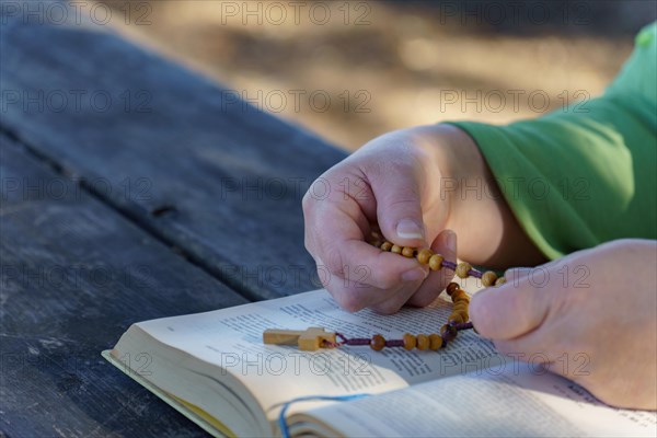 Of a woman's hands praying the rosary beads over a bible in the sunlit countryside