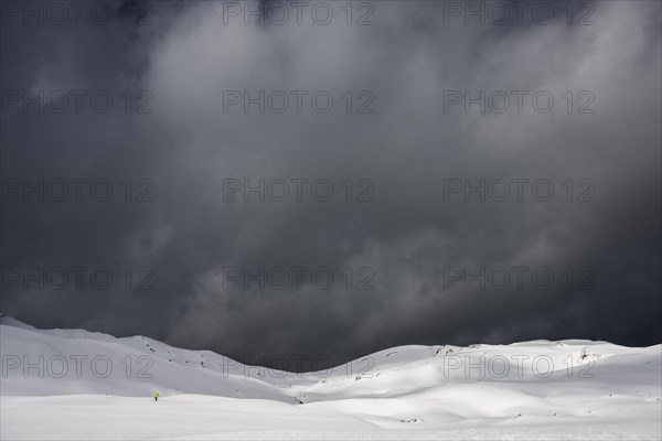 Ski tourer on a vast plateau with snow-covered mountains in the background and dramatic sky