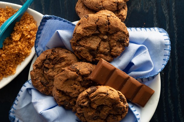 Chocolate chip cookies on a white plate beside brown sugar and a piece of milk chocolate