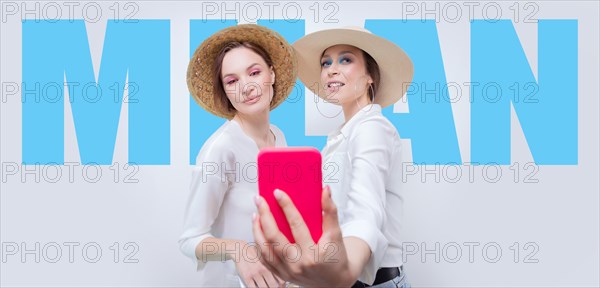 Portrait of two smiling women taking a selfie against the backdrop of a billboard with the inscription Milan . Travel concept.