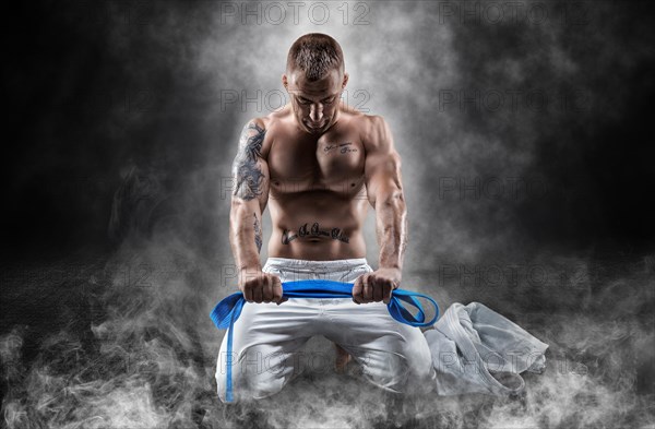 Professional wrestler sits in smoke with a blue belt in his hands and prays. The concept of mixed martial arts