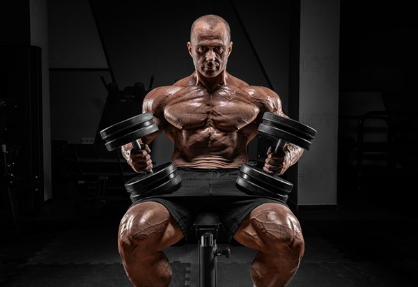 Muscular man sits on a bench with dumbbells. Bodybuilding and powerlifting concept.