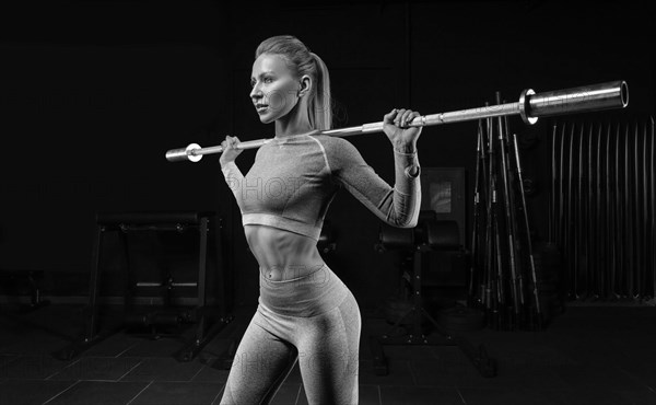 Beautiful tall blonde stands in the gym with a barbell on her shoulders. Squats. Fitness and bodybuilding concept.