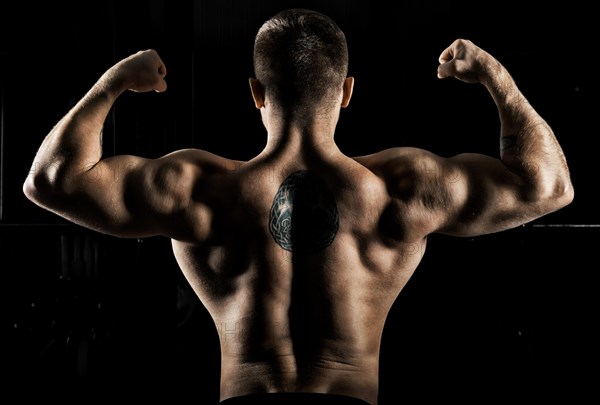 Huge weightlifter is standing with his back to the camera and straining his hands demonstrating a double bicep. Back view