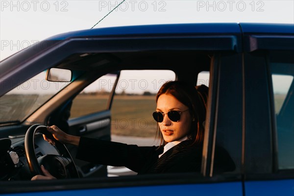 A young beautiful stylish girl driver in a jacket and sunglasses behind the wheel of a car greets passengers