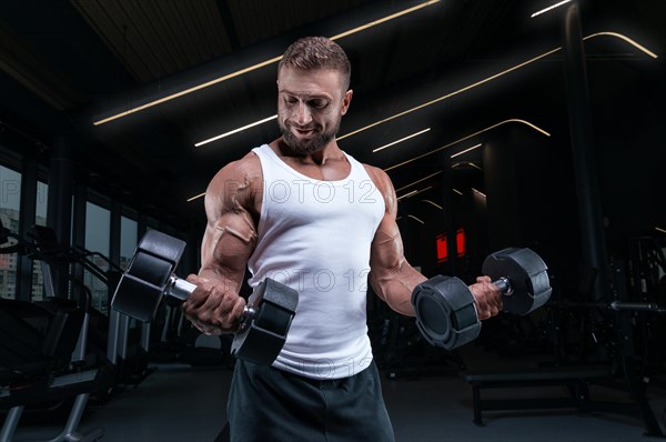 Muscular man in a white t-shirt works out in the gym with dumbbells. Biceps pumping. Fitness and bodybuilding concept.