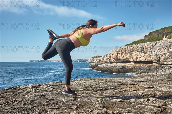 A woman performing a yoga pose on a rocky shore by the sea under a clear sky