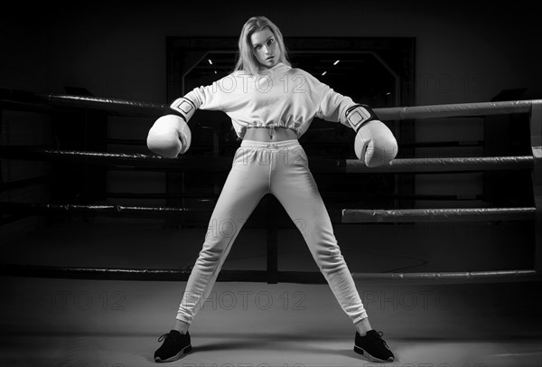 Charming girl in a white sweatshirt poses with huge white gloves against the background of a boxing ring. The concept of sports