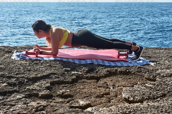 A woman is performing a yoga pose on a mat by the sea on a sunny day