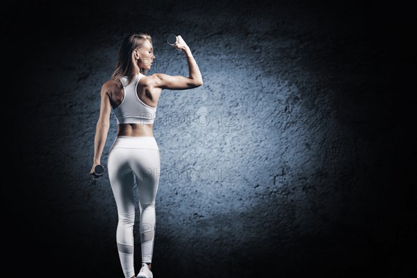 Adorable girl is training in the gym. She lifts the dumbbells up. Back view. The concept of bodybuilding