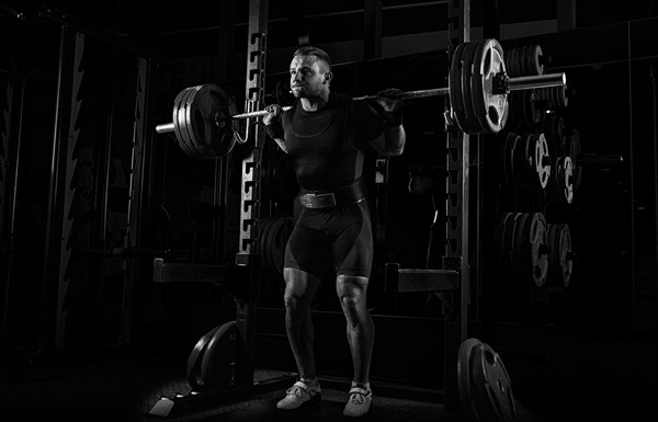 The weightlifter stands with a very heavy barbell on his shoulders. He is at the top and is preparing to sit down