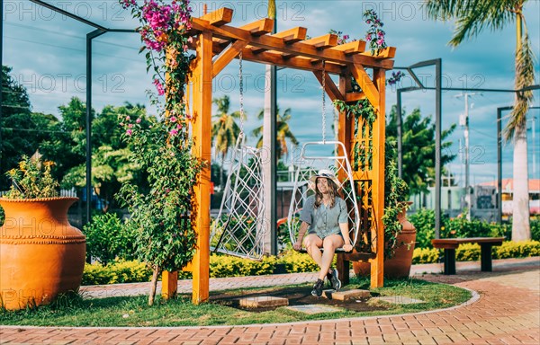 Beautiful girl in hat sitting on a white swing in a beautiful garden. Portrait of a smiling girl sitting on a swing in La Calzada