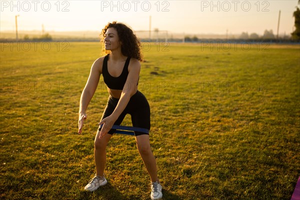 Young curly athletic girl in sportswear performs squats with resistance band outdoors on the grass during sunset