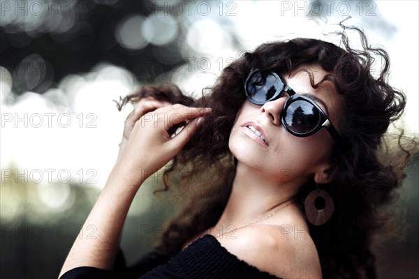 A young slender curly woman in a black dress and sunglasses poses on a sunny day in a city park