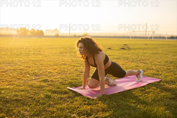 Young curly athletic girl in sportswear doing leg stretching on a yoga mat outdoors on the grass during sunset