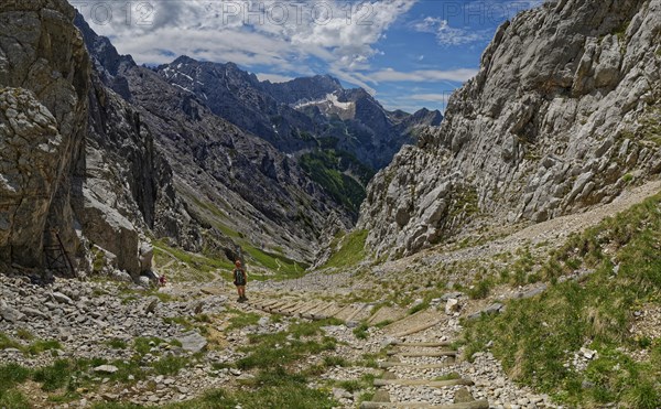 A mountaineer walks down a stony path in a mountainous valley under a cloudy sky