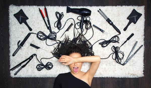 The delightful girl barber covered her face with her hand and stuck out her tongue. On the background there are accessories for haircuts and hairdressing