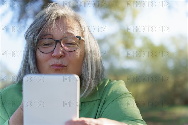 Mature white-haired woman with glasses blowing a kiss to her digital tablet in a video call in the countryside