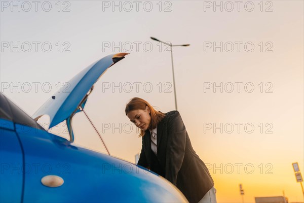 A young beautiful stylish girl driver in a jacket is trying to identify the cause of a car breakdown under the hood