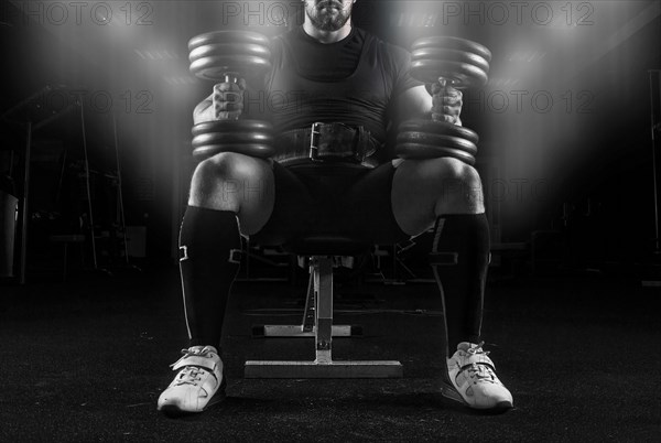 Professional weightlifter sits on a bench in the gym with two dumbbells on his lap