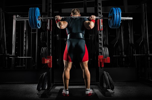 Professional athlete is standing with a barbell on his shoulders and is about to sit down with her. View from the back