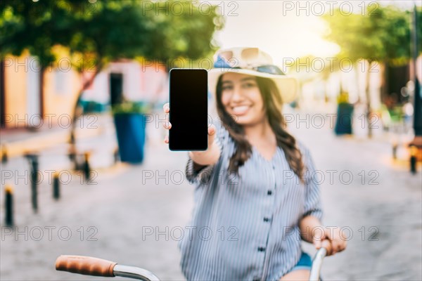 Smiling tourist on bicycle showing cell phone screen on the street. Beautiful girl with hat on bicycle showing cell phone screen on the street