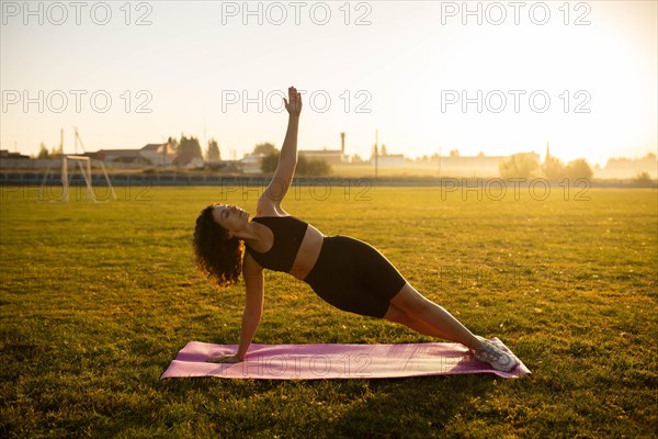 Young curly athletic girl in sportswear performs a side plank exercise on a yoga mat outdoors on the grass during sunset