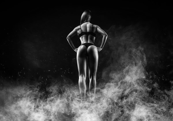Image of a female athlete in a fire arena surrounded by smoke. Back view. Bodybuilding and fitness concept.