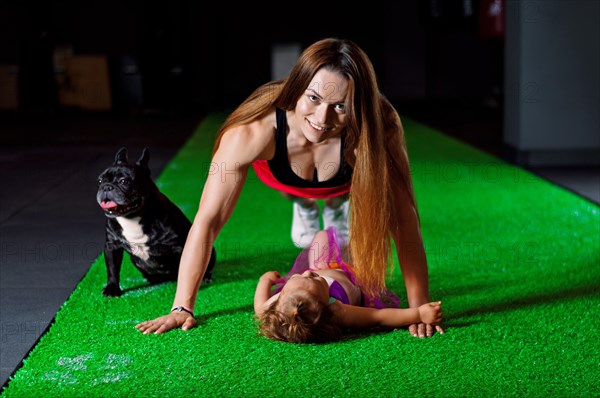 Charming sports mom plays in the gym with her little daughter and a French bulldog.