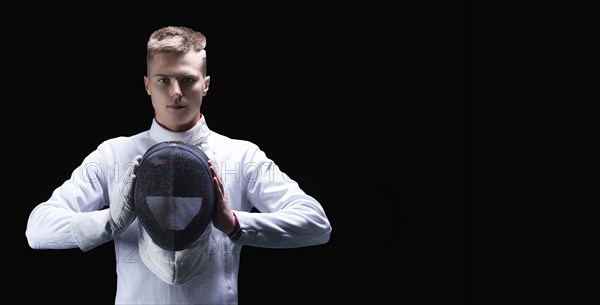 Portrait of a male fencer on a black background. He holds a helmet in front of him. The concept of fencing.