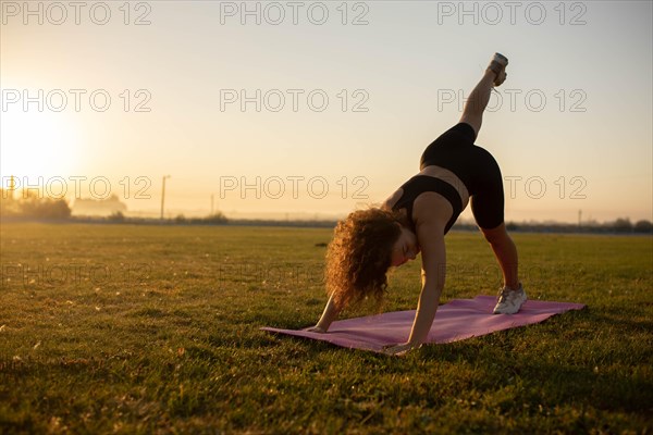 Young curly athletic girl in sportswear doing yoga on a yoga mat outdoors on the grass during sunset