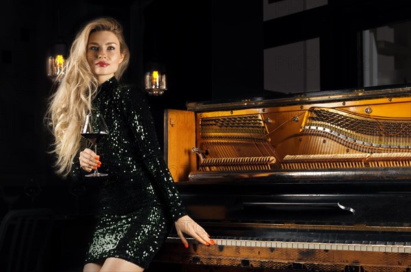 Charming girl in a green evening dress is smiling and holding a glass of wine in her hand while sitting on a retro piano.