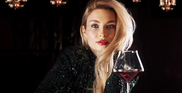 Seductive blonde in a green evening dress posing with a glass of red wine.