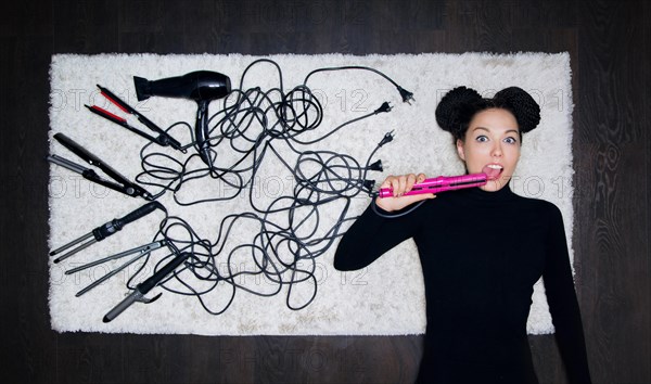 Young charming girl with dreadlocks on her head pressed her tongue into the hair iron and looked at the camera in amazement. The girl is lying on a white carpet. Around tools for creating haircuts