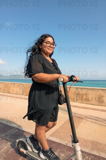 A joyful woman rides an electric scooter by the seaside on a clear sunny day