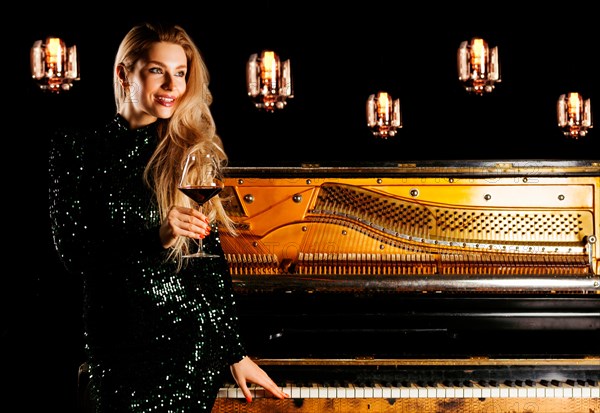 Charming girl in evening dress sits on the piano and poses for the camera with a glass of wine.