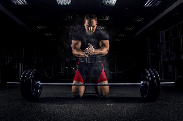 Ahlete kneels in the gym before the bar and claps his hands after which the dust from the chalk flies