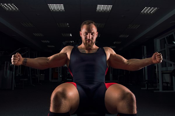 The weightlifter sits on a bench in the gym with arms outstretched and smiles