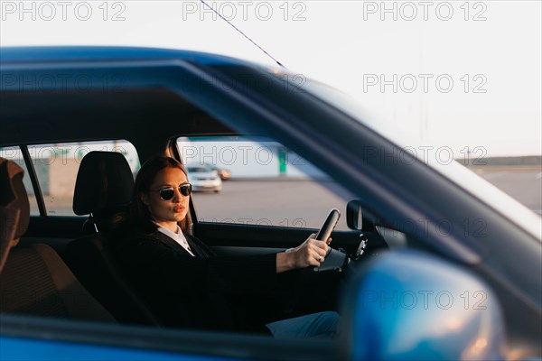 A young beautiful stylish girl driver in a jacket and sunglasses at the wheel of a car is waiting for passengers