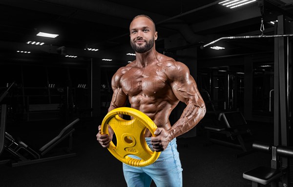 Pumped-up man posing in the gym in jeans with a dumbbell in his hand. Sports