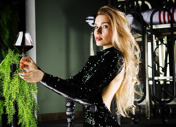 Seductive blonde in a green evening dress posing with a glass of red wine.